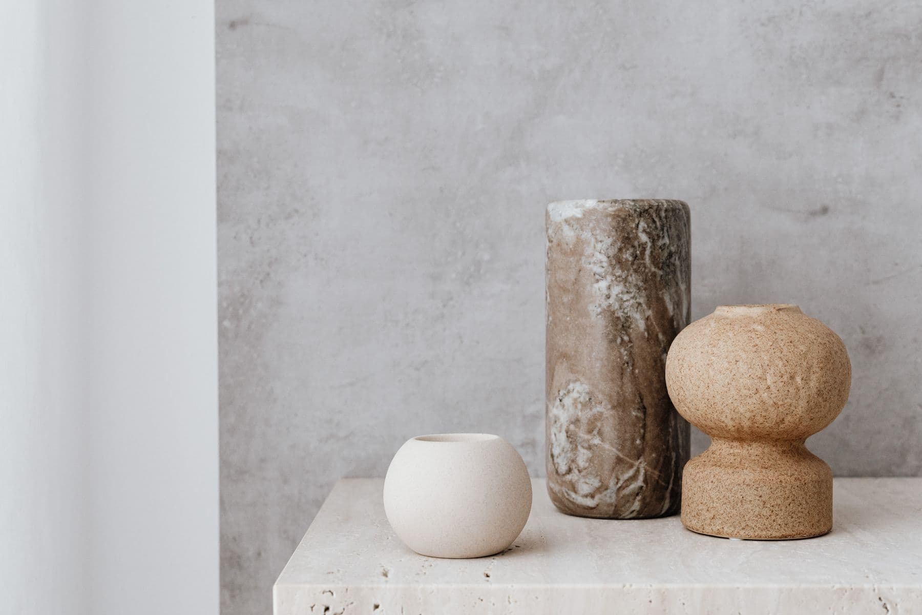 Three stone and ceramic vessels in warm neutral tones on a travertine plinth